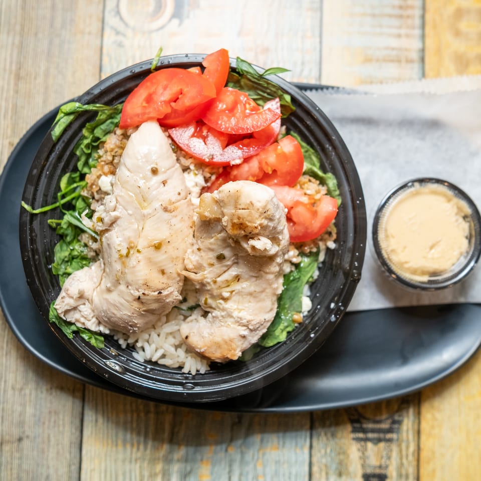 Chicken breast, rice, quinoa, spinach, and tomatoes in a black bowl with a side of sauce.