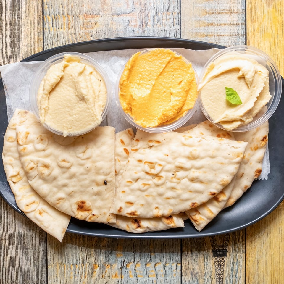 Three distinct hummus dips and flatbread on a dark plate, set on a wooden table.