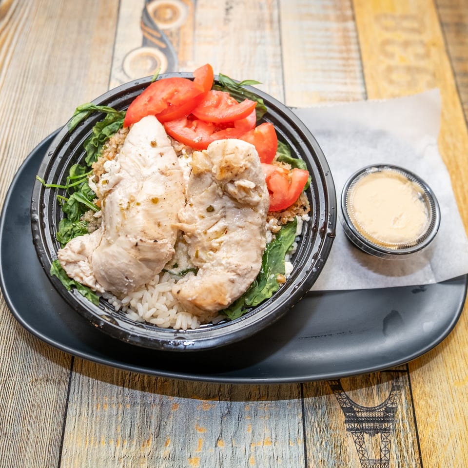 Chicken breast, rice, tomatoes, and greens in a black bowl with a side of creamy sauce.