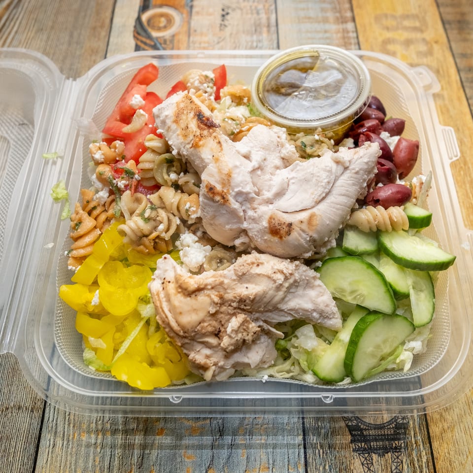 Grilled chicken salad with pasta, tomatoes, feta, olives, cucumbers, and peppers in a plastic container.