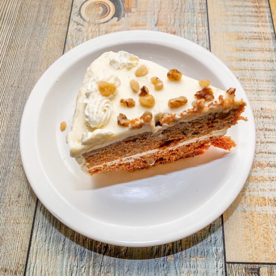 Slice of carrot cake with cream cheese frosting and walnuts on a white plate.